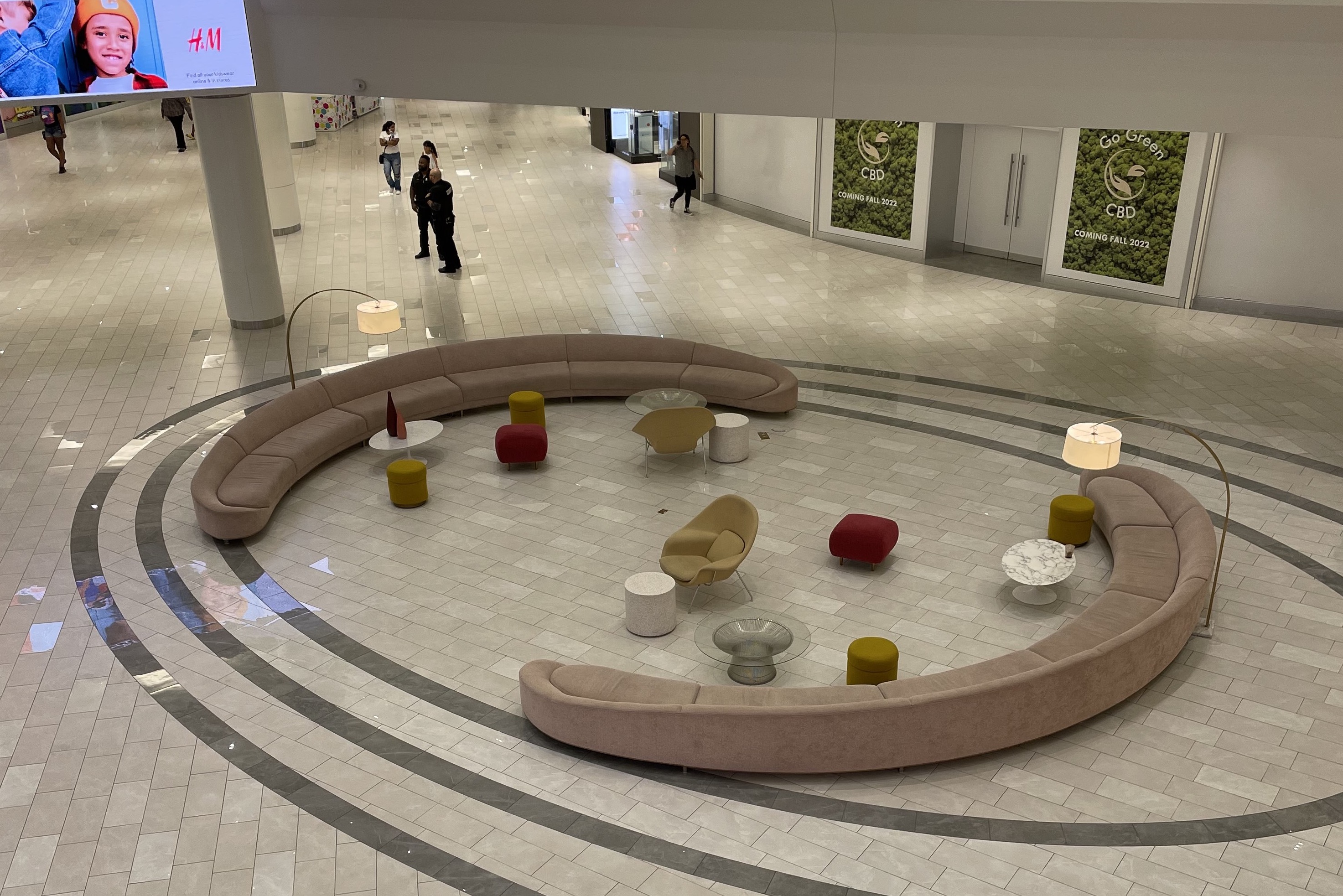 Seen from above, a pastel set of plush furniture—two long, rounded couches and a smattering of chairs and ottomans—dominates the center of a court in what looks to be an indoor retail space with a tiled floor.